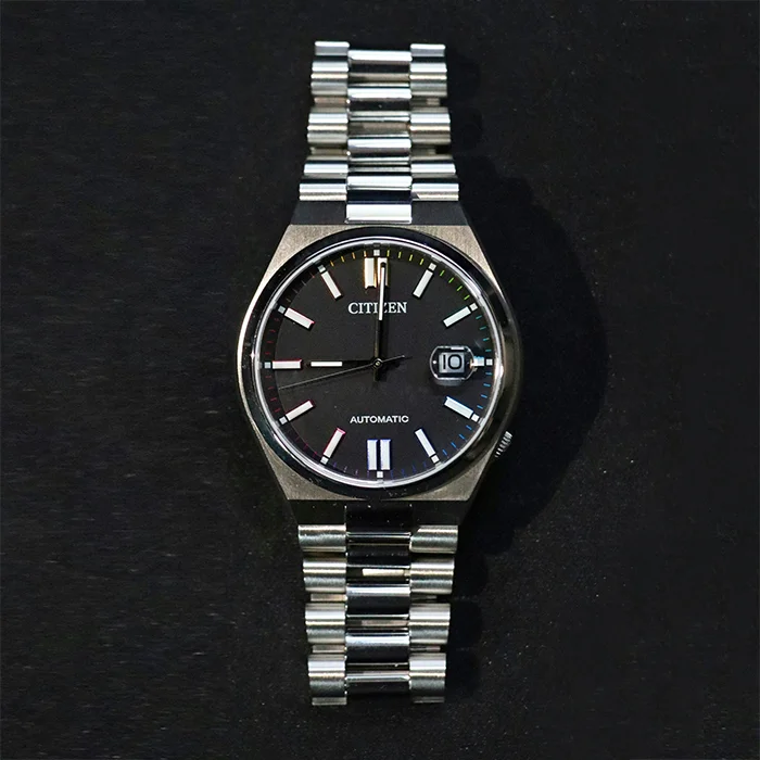 Unedited photo of a steel automatic watch with a black dial, shot on a dark textured background with dust